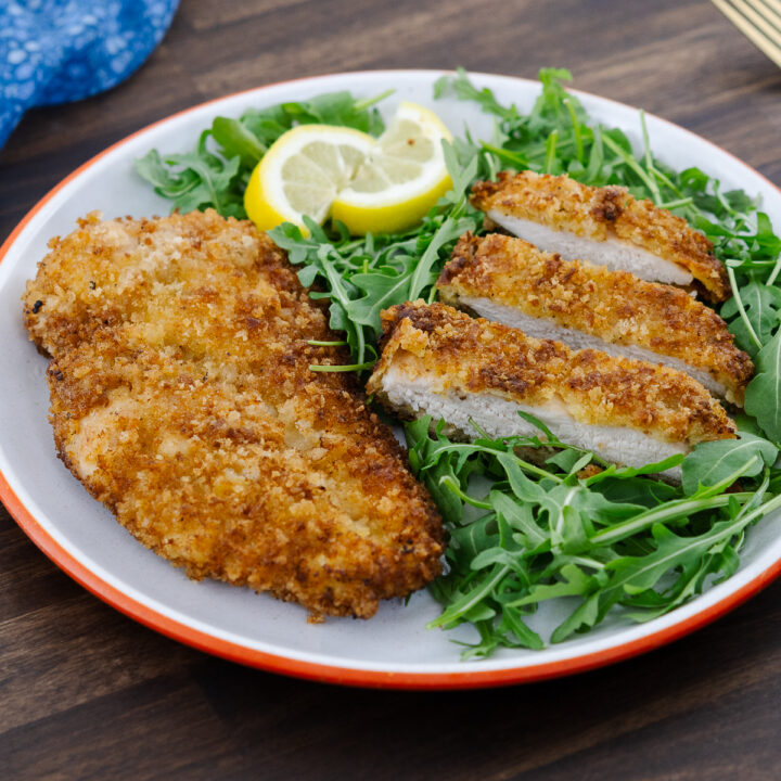A crispy chicken schnitzel served on a plate with baby arugula as a side.