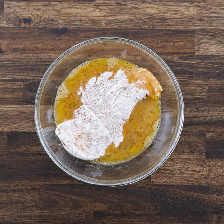A piece of flour-coated chicken breast being dipped into the egg mixture in a bowl.