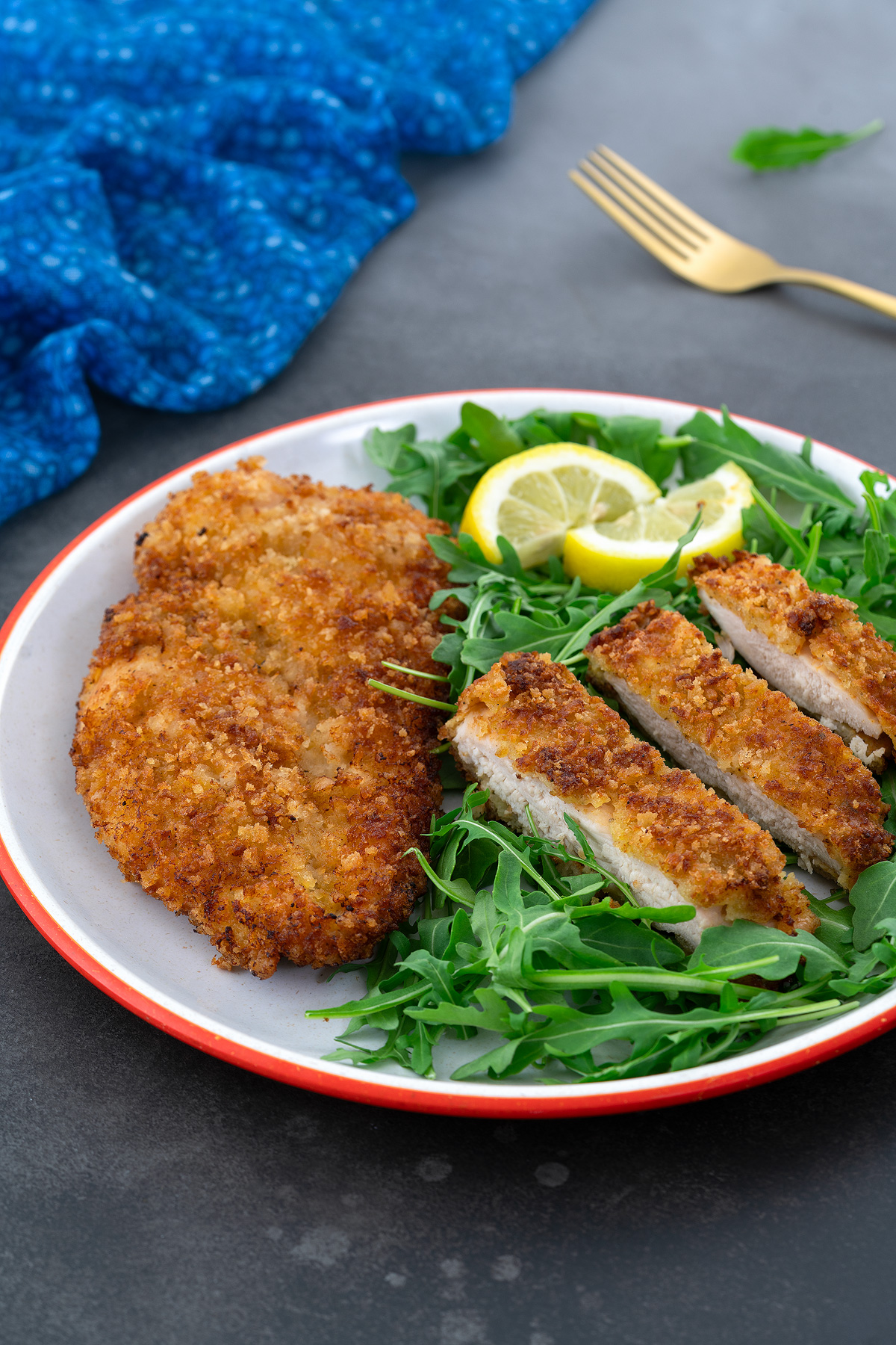 Whole and sliced chicken breast schnitzel on a white plate, served with baby arugula. A golden fork and blue towel are placed beside the plate.