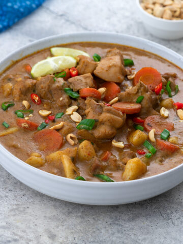 Thai Massaman Curry in a white bowl on a white table, surrounded by a cup of peanuts, chopped spring onions, and a blue towel.