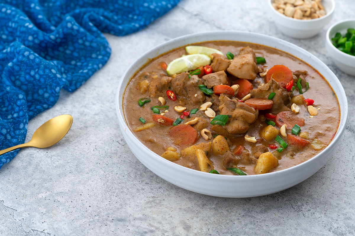 Thai Massaman Curry in a white bowl on a white table, surrounded by a cup of peanuts, chopped spring onions, a blue towel, and a golden spoon.