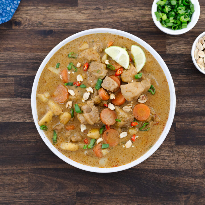 Bowl of Massaman curry topped with peanuts, chili, lime wedges, and spring onions.