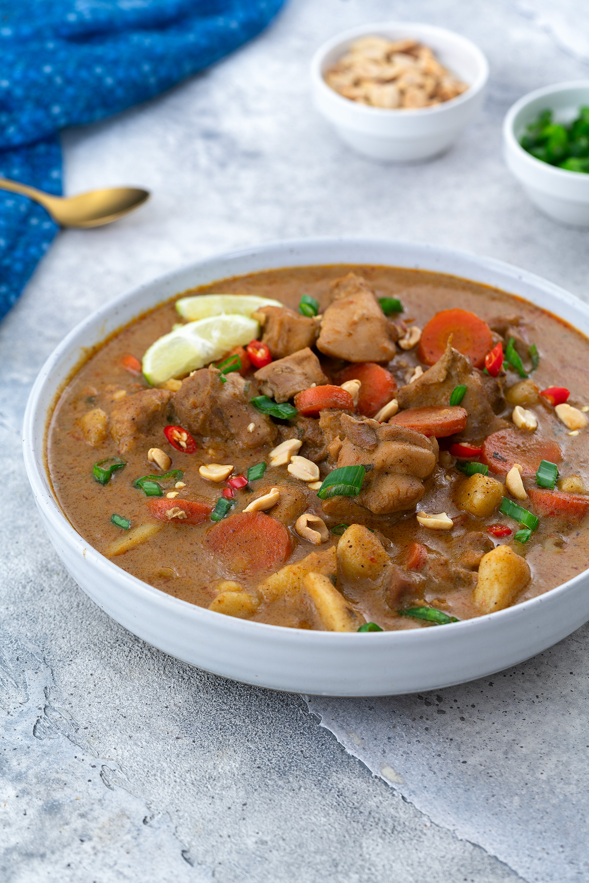 Thai Massaman Curry in a white bowl on a white table, surrounded by a cup of peanuts, chopped spring onions, a blue towel, and a golden spoon.