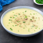 A bowl of potato leek soup on a gray table, surrounded by a blue towel,a small cup of chives.