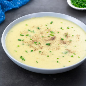 A bowl of potato leek soup on a gray table, surrounded by a blue towel,a small cup of chives.