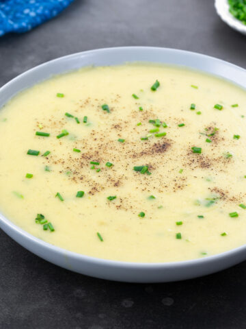 A bowl of potato leek soup on a gray table, surrounded by a blue towel,a small cup of chives.