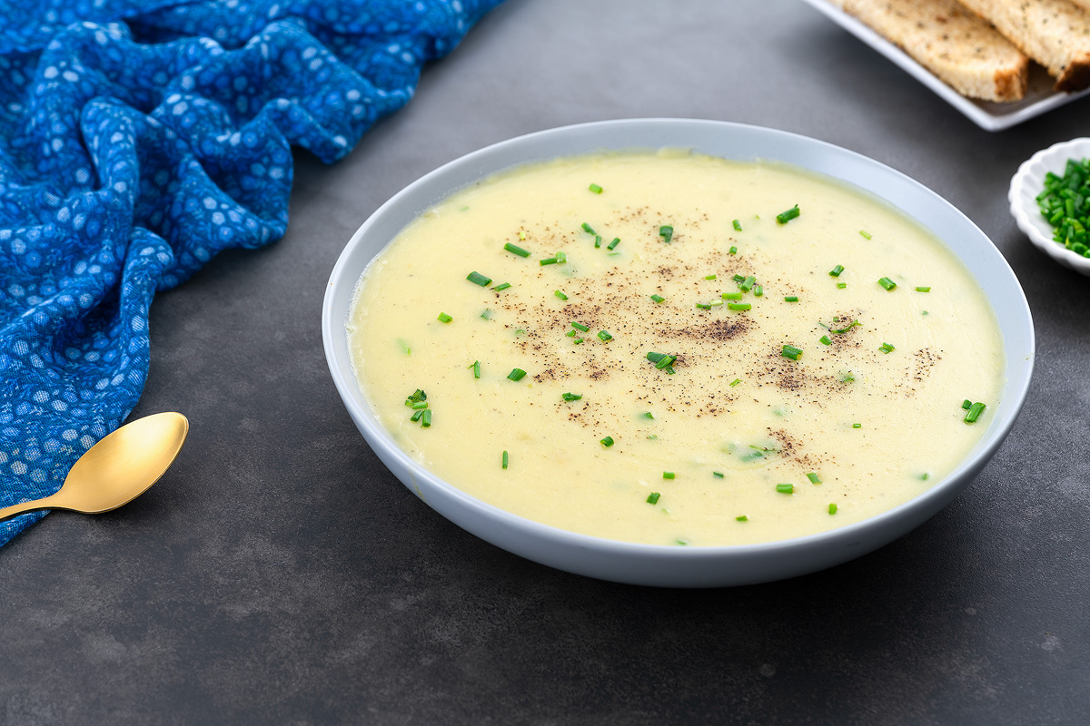 A bowl of potato leek soup on a gray table, surrounded by a blue towel, a golden fork, a small cup of chives, and a few pieces of bread.