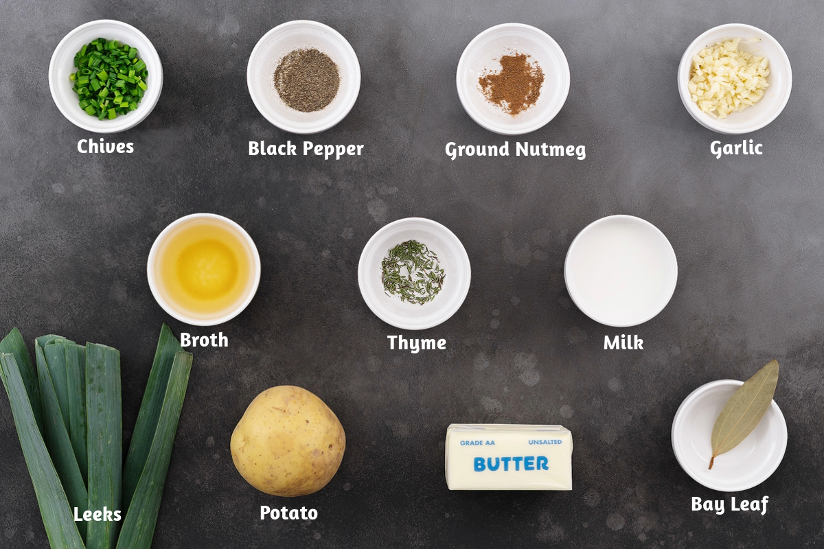 Potato leek soup ingredients arranged on a gray table, including chives, black pepper, nutmeg, garlic, broth, thyme, milk, leeks, potatoes, butter, and a bay leaf.