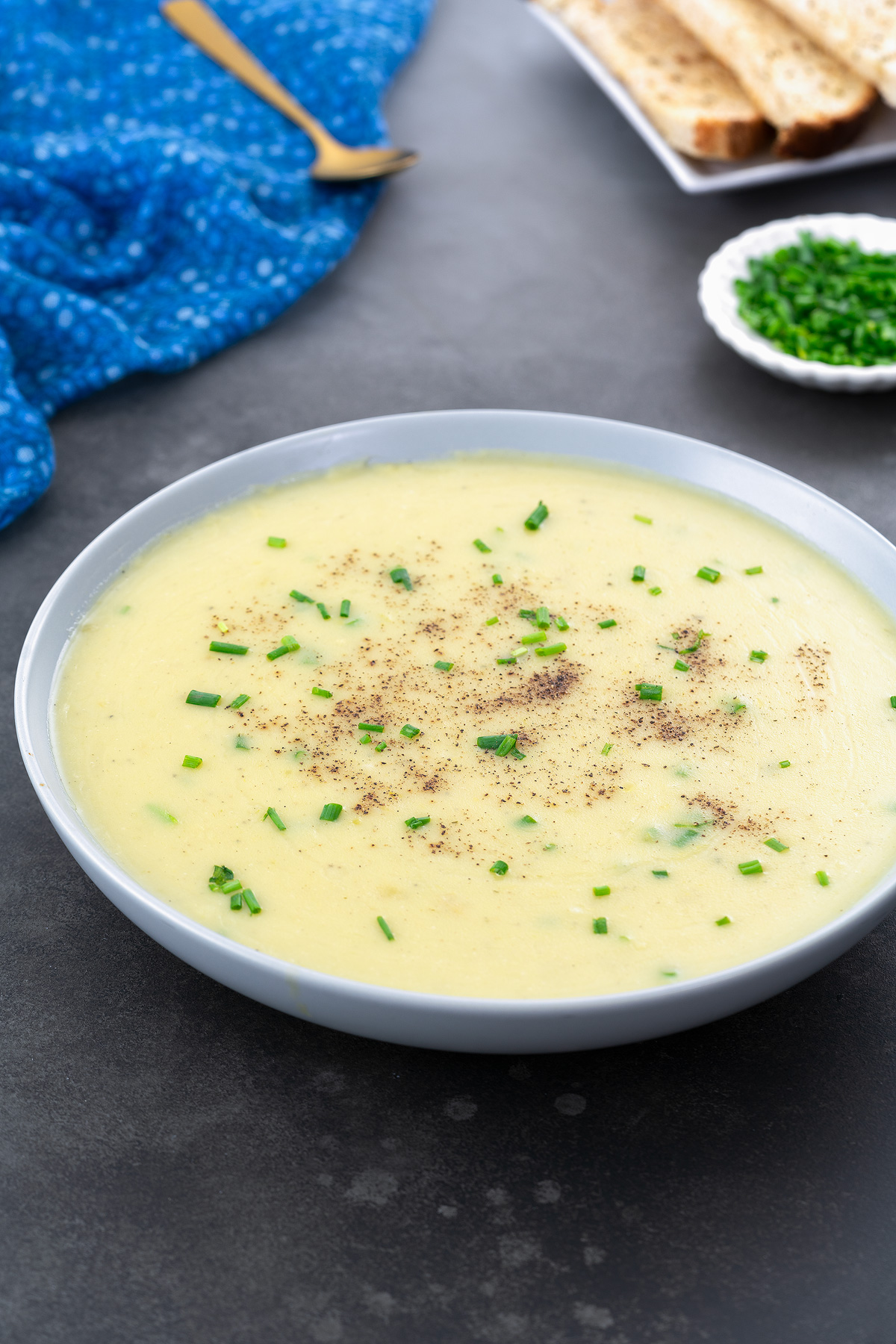 A bowl of potato leek soup on a gray table, surrounded by a blue towel, a golden fork, a small cup of chives, and a few pieces of bread.