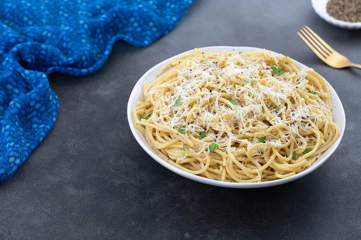 A white cup of Cacio e Pepe on a gray table, with a small plate of ground black pepper, a golden fork, and a blue towel nearby.