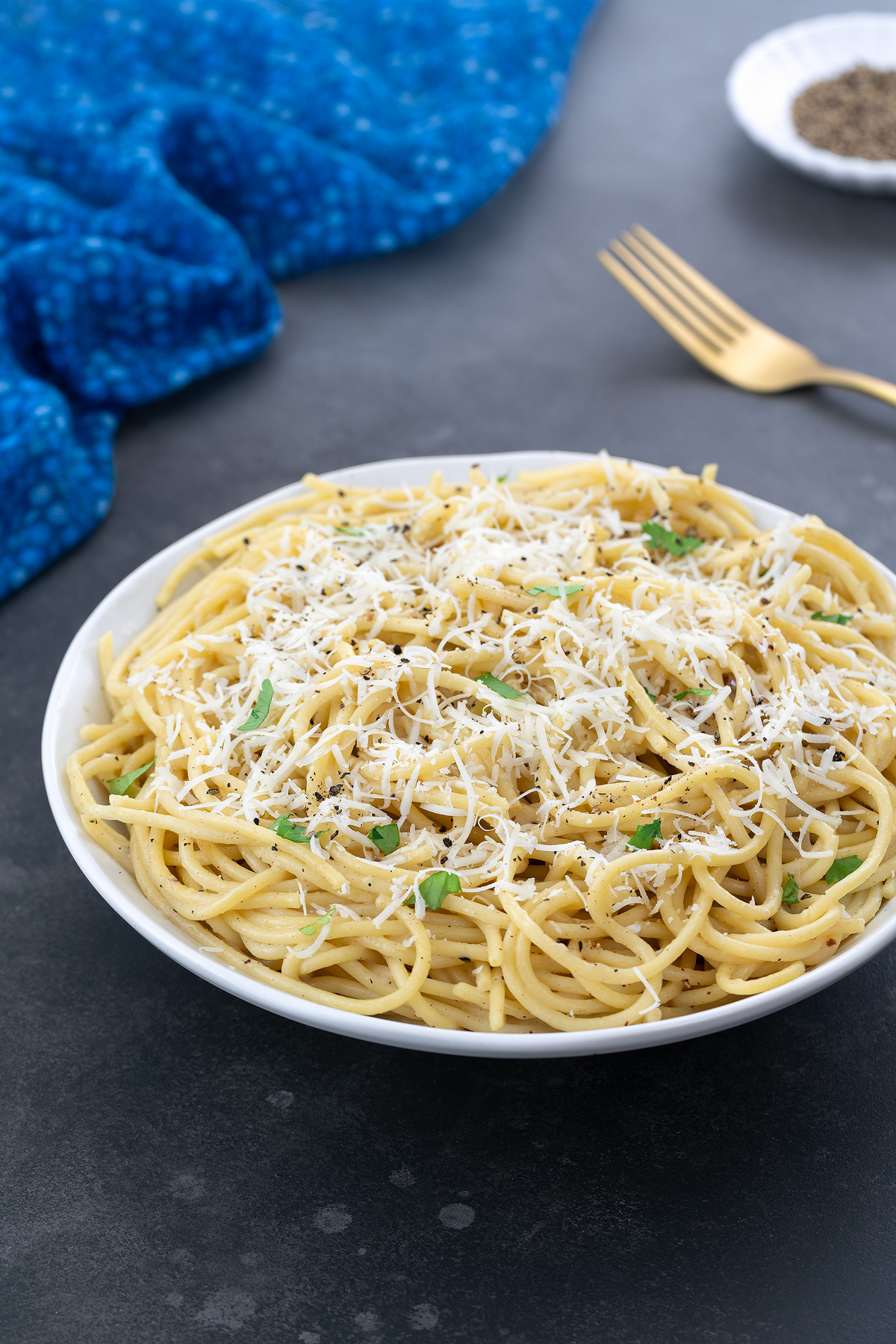 A white cup of Cacio e Pepe on a gray table, with a small plate of ground black pepper, a golden fork, and a blue towel nearby.