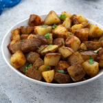 A white bowl of homemade fried potatoes on a white table, with a blue towel next to it.