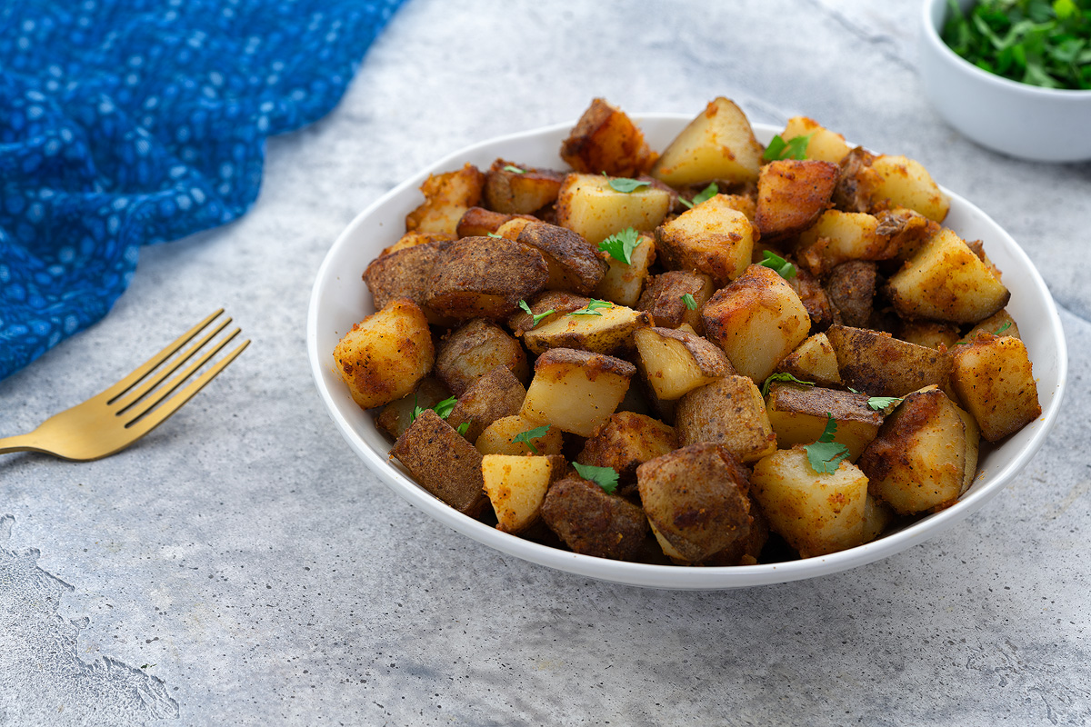 A white bowl of homemade fried potatoes on a white table, with a cup of cilantro, a golden fork, and a blue towel beside it.