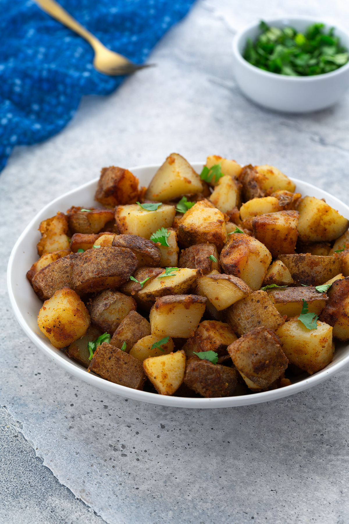 A white bowl of homemade fried potatoes on a white table, with a cup of cilantro, a golden fork, and a blue towel beside it.