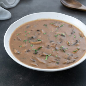 A white bowl of mushroom gravy placed on a gray table, with a cream-colored towel, and a wooden spoon arranged around it.