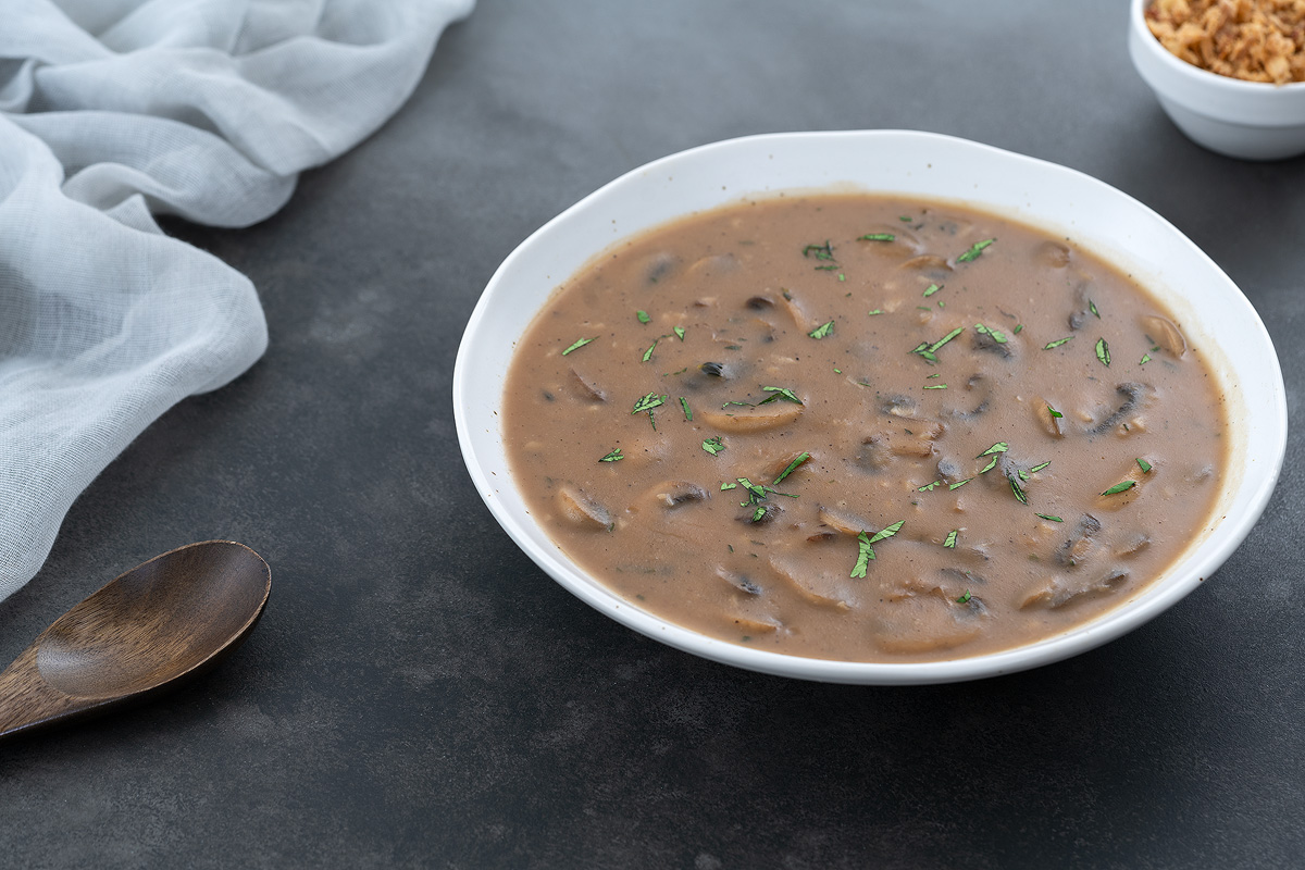 A white bowl of mushroom gravy placed on a gray table, with a cream-colored towel, a small cup of fried onions, and a wooden spoon arranged around it.