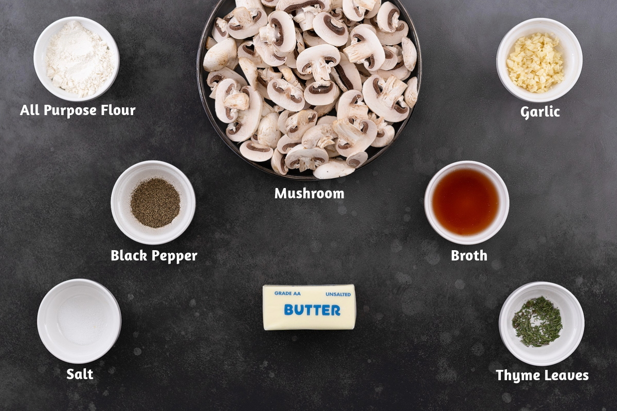 Mushroom gravy ingredients placed on a gray table, including all-purpose flour, mushrooms, garlic, black pepper, broth, salt, butter, and thyme leaves.