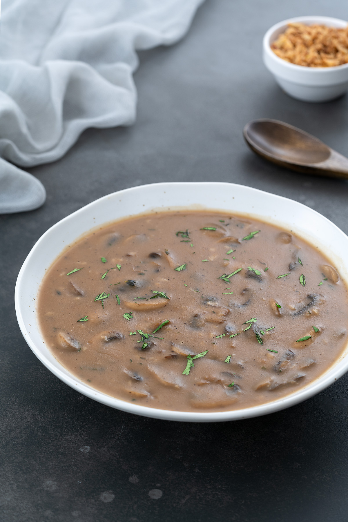 A white bowl of mushroom gravy placed on a gray table, with a cream-colored towel, a small cup of fried onions, and a wooden spoon arranged around it.