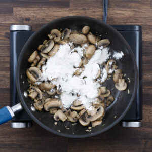 Sautéed mushrooms in a pan being sprinkled with flour.