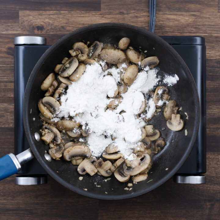 Sautéed mushrooms in a pan being sprinkled with flour.