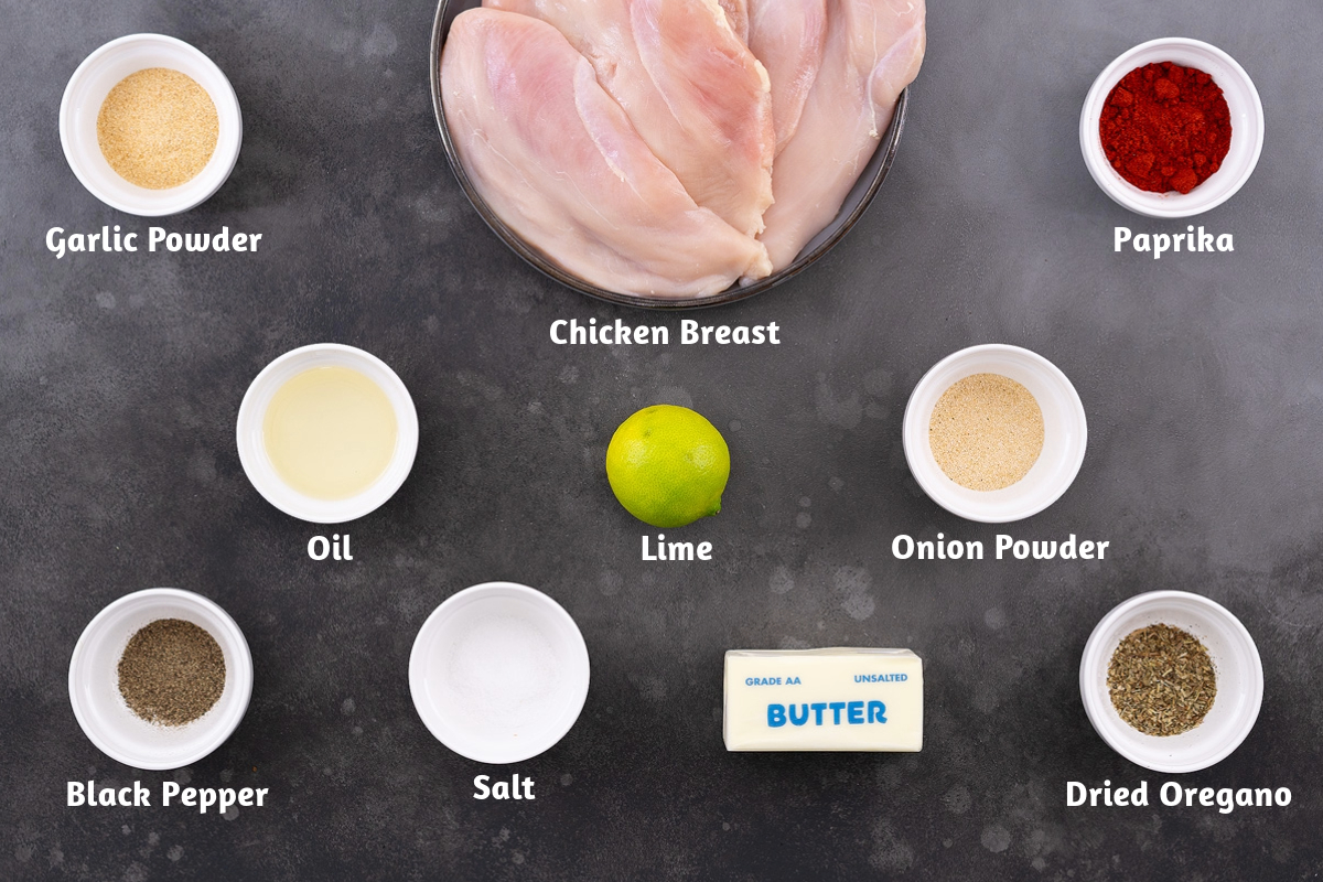 Ingredients for pan-seared chicken breast recipe arranged on a gray table, including garlic powder, chicken breast, paprika, oil, lime, onion powder, black pepper, salt, butter, and dried oregano.