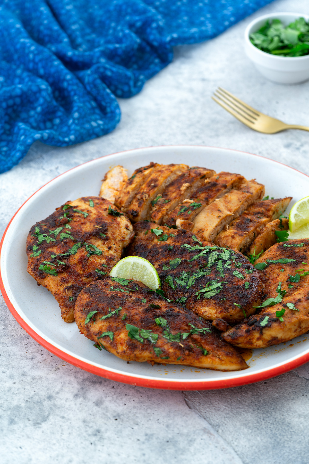 Pan-seared chicken breast with sliced pieces arranged on a plate, surrounded by a blue towel, a golden fork, and a cup of cilantro.